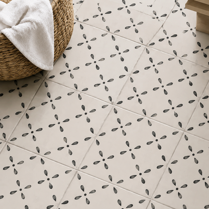 Beige ceramic floor tiles with a repeating black geometric cross pattern, styled in a bathroom setting with a woven basket and white towel in the corner.